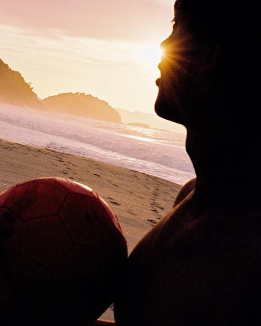 Silhouette of shirtless man on beach holding soccer ball at sunset with lens flare, ocean, sand, and landscape backdrop.