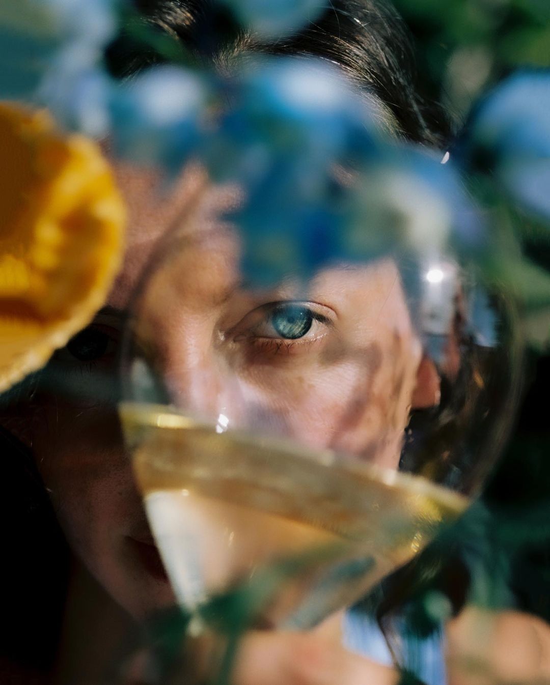 Close-up portrait of an adult female with blue eyes, partially visible through a glass of amber liquid and blurred yellow and blue flowers.