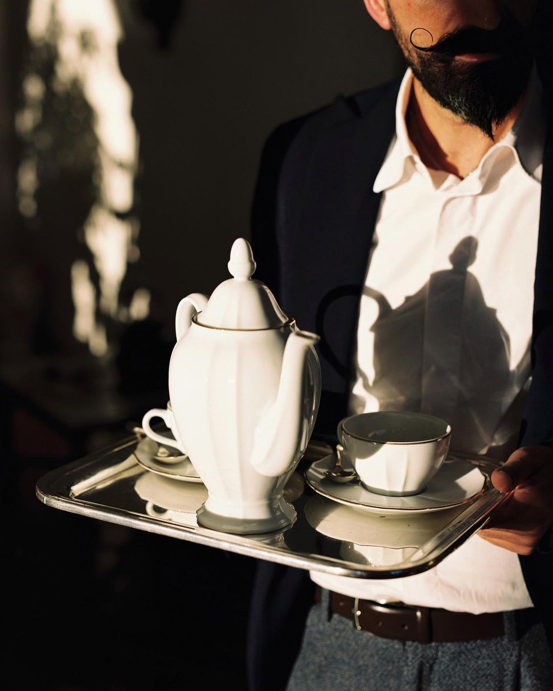 Man with mustache holds a silver tray with a white teapot and cups. The image is well-lit with shadows and rich with detail.