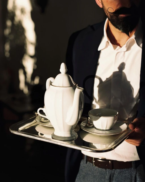 Man with mustache holds a silver tray with a white teapot and cups. The image is well-lit with shadows and rich with detail.