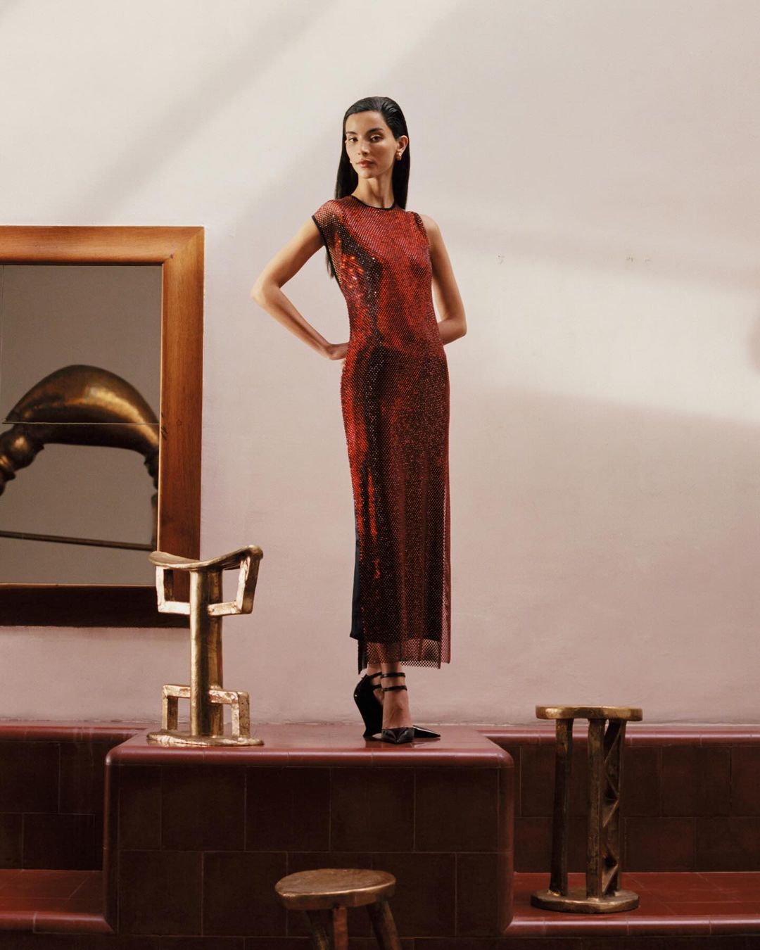 Fashionable woman posing in red gown with elegant gold furniture in minimal interior setting with warm lighting and clean composition.