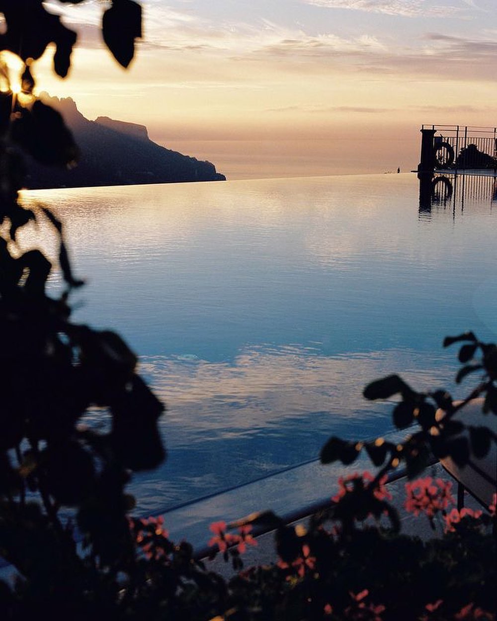 Scenic view of an infinity pool overlooking the ocean with mountains in the background during a vibrant sunset with pink and blue sky.