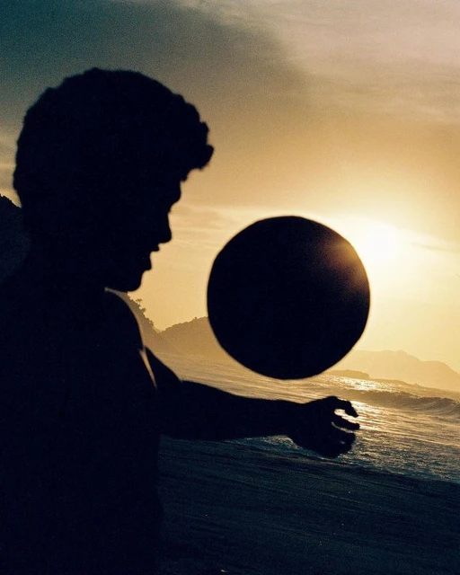 Silhouette of a person holding a ball on a beach at sunset with golden light, ocean waves, and distant land in the background.