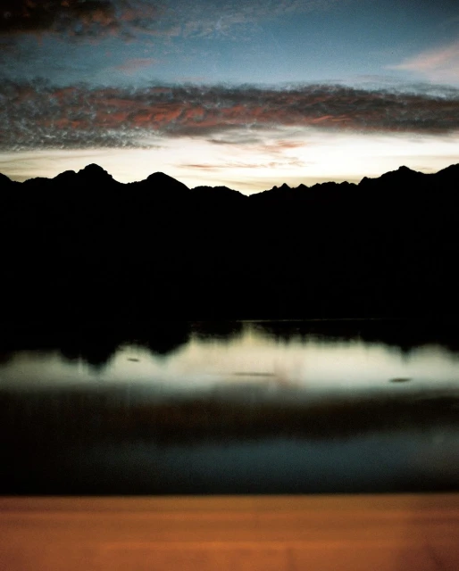 Silhouette of mountains reflecting in a serene lake at sunset under a sky filled with dramatic clouds in a horizontal, landscape shot.