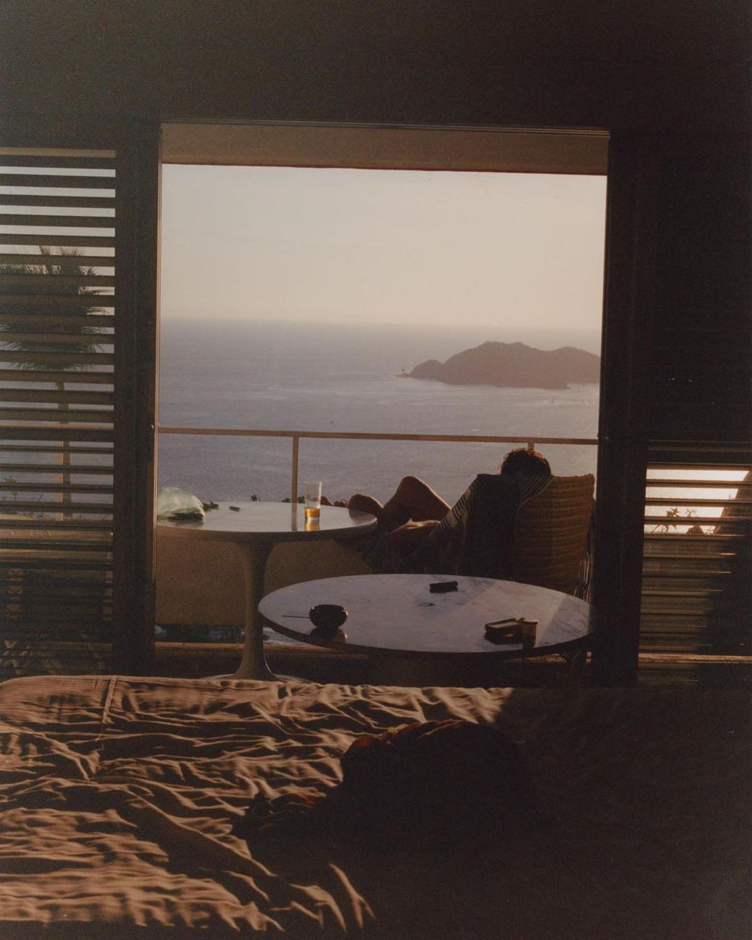 Person relaxing on a balcony with ocean view. Features a table with a bottle, visible from a bedroom interior with neutral tones.