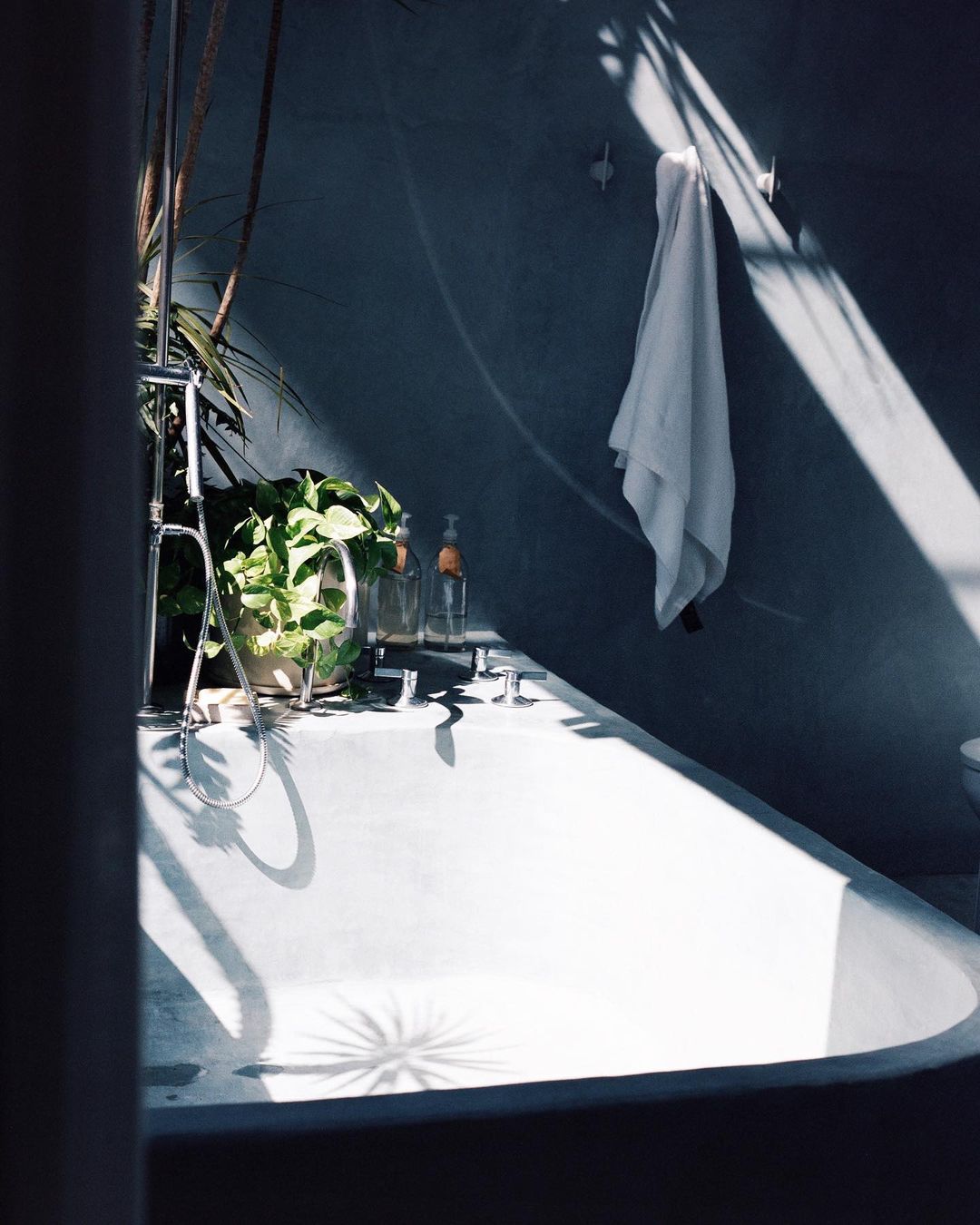 Bathtub with sunlight and plants creating shadows, in a grey bathroom with a white towel hanging on the wall.