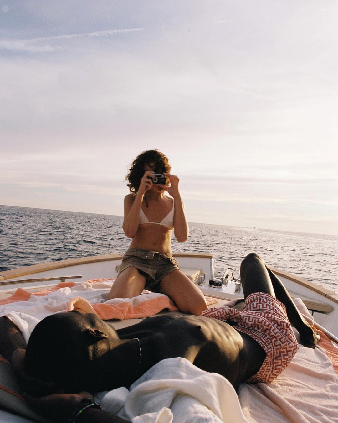 Two people relaxing on a boat in the sunshine: a woman takes a photo while kneeling, and a man lies down on a blanket, shirtless.