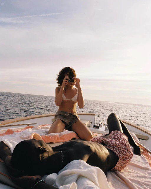 Two people relaxing on a boat in the sunshine: a woman takes a photo while kneeling, and a man lies down on a blanket, shirtless.