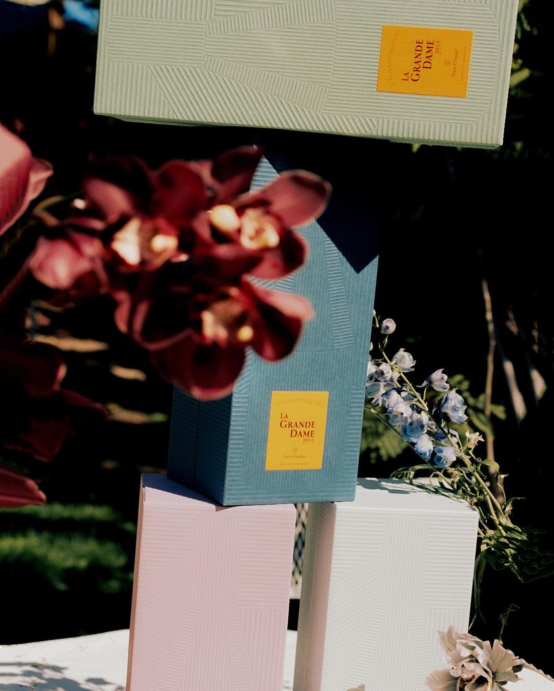 Stacked La Grande Dame champagne boxes with floral accents in a sunlit, outdoor setting, capturing luxury and elegance in an artistic still life.