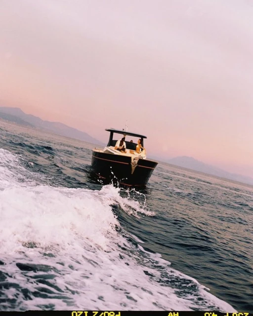 Two people on a wooden motorboat creating waves on blue water with pinkish sky and distant mountains in the background on a sunny day.