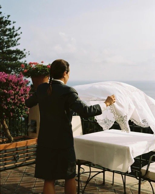 A woman in a blazer sets a white tablecloth on an outdoor table on a terrace with a potted plant on the railing and an ocean view.