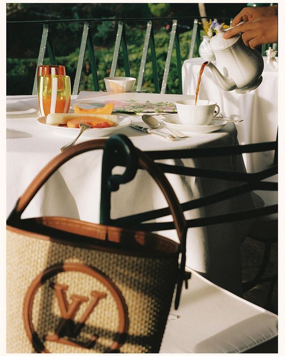 Tea pouring into a cup with fruit plate and Louis Vuitton bag on table outdoors, set on balcony, creating a scene of leisure.