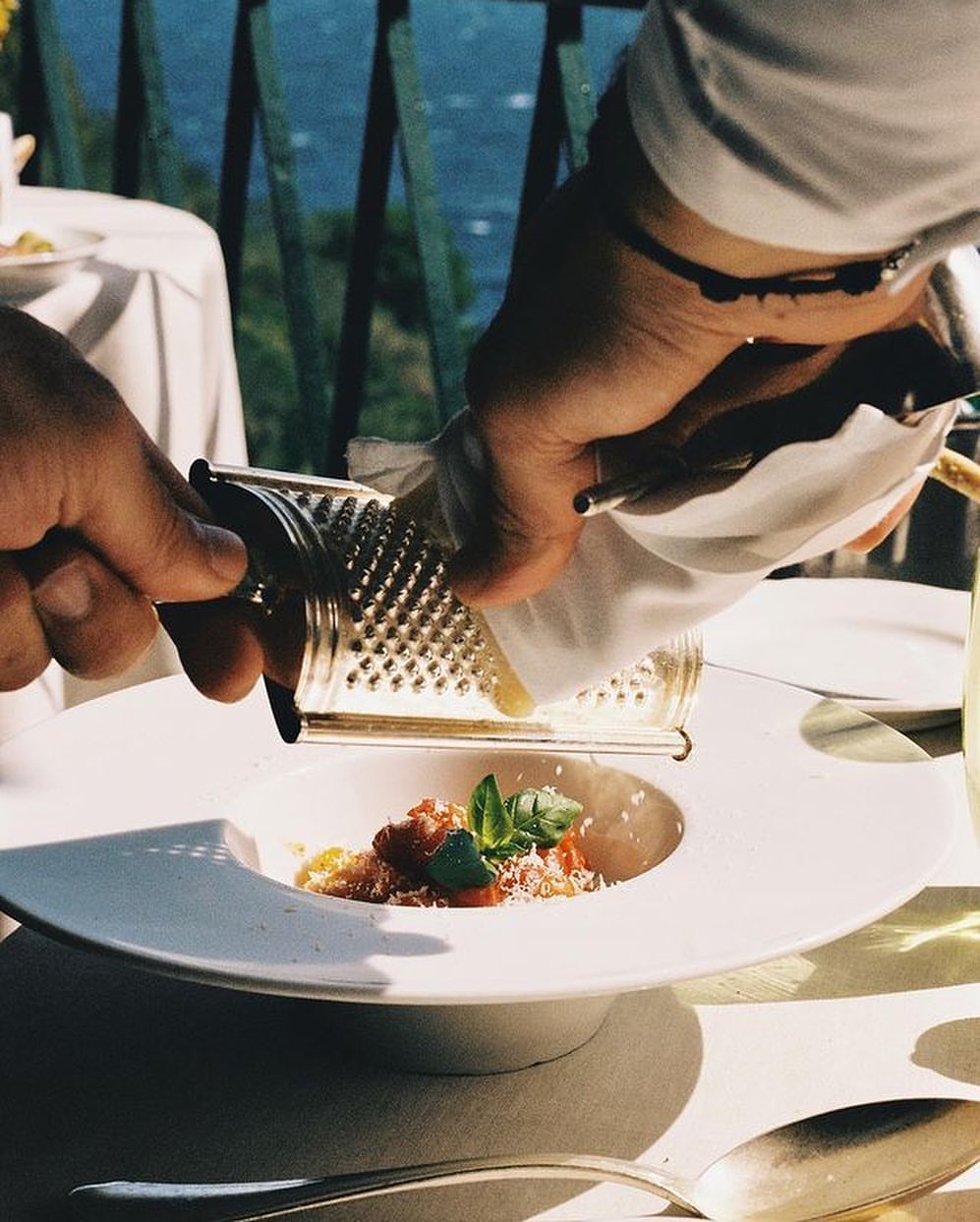 Overhead shot of a person grating cheese over a pasta dish on a white plate at a bright table on a sunny day, outdoor setting.