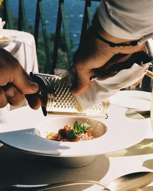 Overhead shot of a person grating cheese over a pasta dish on a white plate at a bright table on a sunny day, outdoor setting.