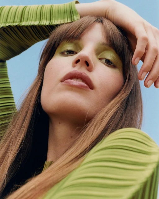 Close-up portrait of a woman with green eyeshadow and a green textured shirt against a bright blue sky, shot from a low angle.