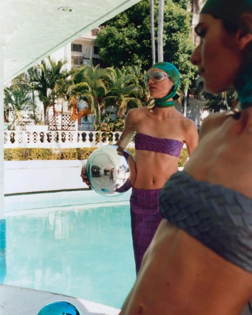 Two women in headscarves and coordinated outfits pose near a turquoise pool with palm trees and architecture in the sunny background.