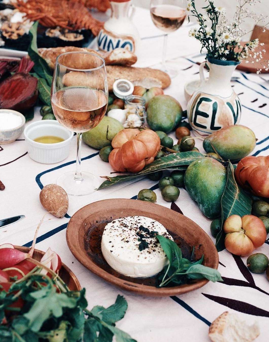 Overhead view of a table setting with various fruits, vegetables, wine, cheese, and bread arranged on a white patterned tablecloth.