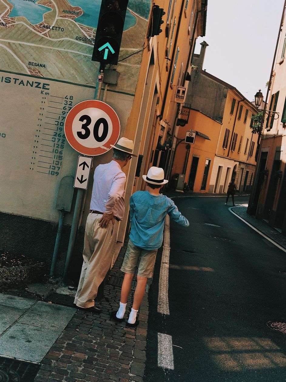 An adult male and child stand on a city street next to a sign with numbers, both wearing hats and casual clothes on a sunny day.
