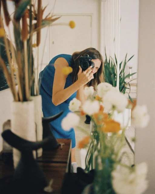 Woman in blue dress photographs reflection with camera, surrounded by flowers and interior decor in a naturally lit indoor space.