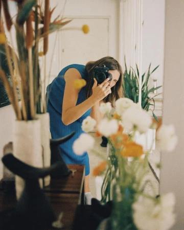 Woman in blue dress photographs reflection with camera, surrounded by flowers and interior decor in a naturally lit indoor space.