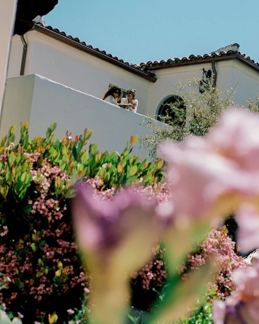 Two children stand on a sunlit balcony of a white building under a clear blue sky, framed by greenery and pink flowers in the foreground.