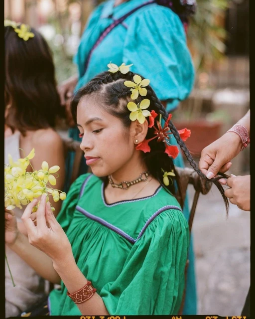 A woman in a green dress with flowers in her braided hair holds a flower, sitting outside, while someone braids her hair.