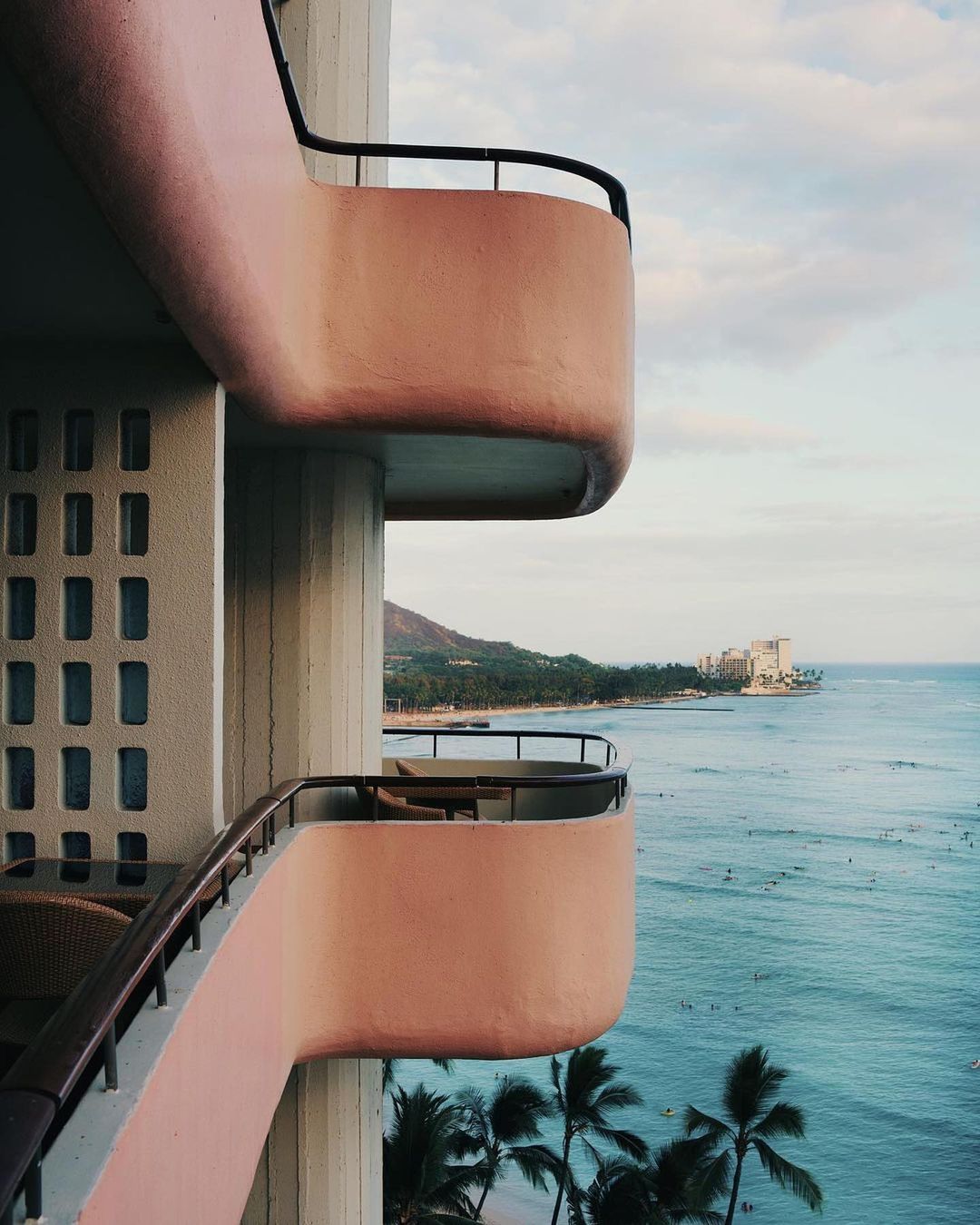 Coastal hotel balconies offer scenic views of the ocean with surfers, palm trees, and a distant mountain under a blue and cloudy sky.
