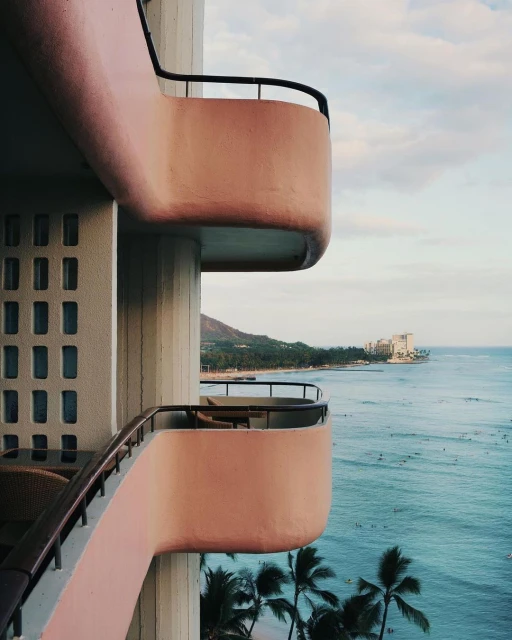 Coastal hotel balconies offer scenic views of the ocean with surfers, palm trees, and a distant mountain under a blue and cloudy sky.