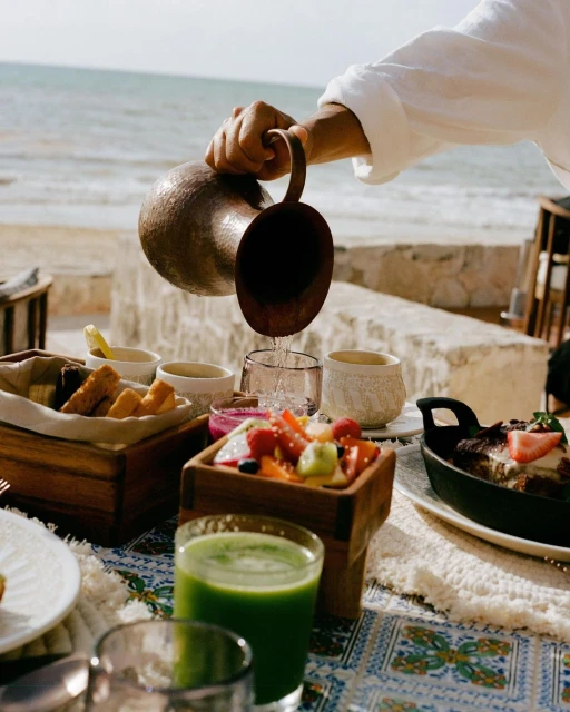 Person pours water from copper pitcher into glass at outdoor table with fresh juice, fruit, and bread, with an ocean view.