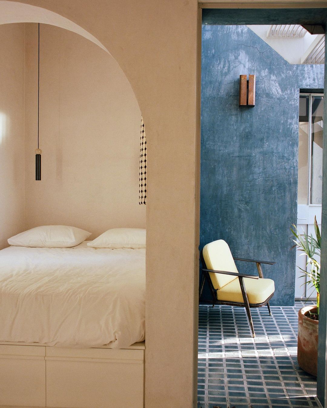 Minimalist interior featuring a white bed in the foreground and an adjacent outdoor patio with a yellow chair in the background.