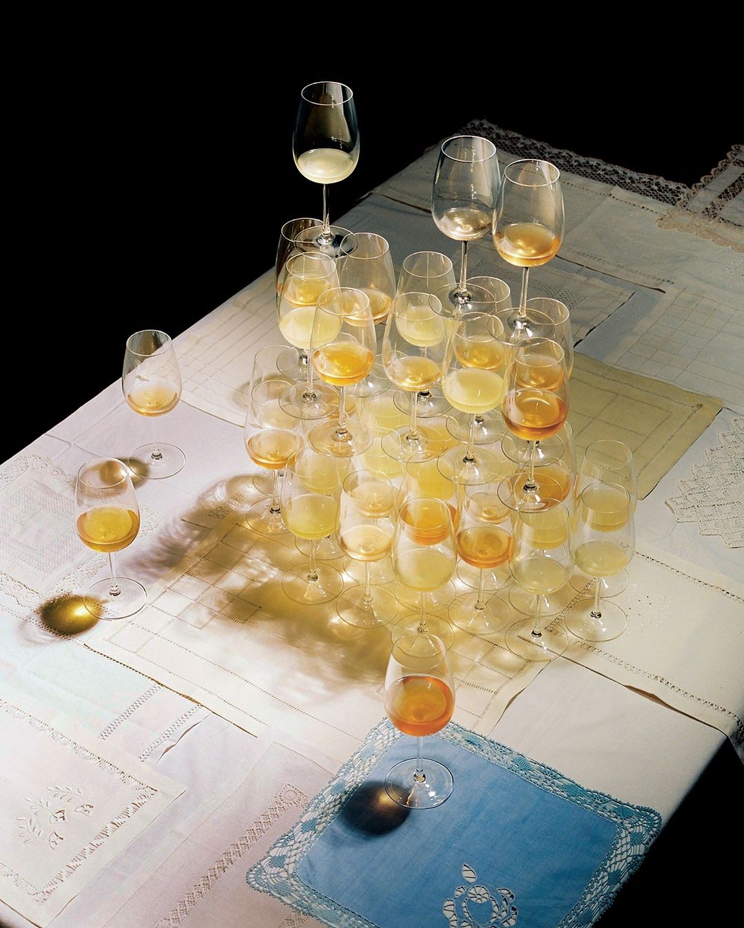 A pyramid of wine glasses, some filled with liquid, sits on a tablecloth against a dark background, creating a complex play of light.