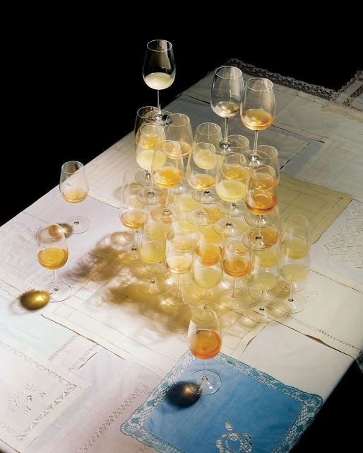 A pyramid of wine glasses, some filled with liquid, sits on a tablecloth against a dark background, creating a complex play of light.