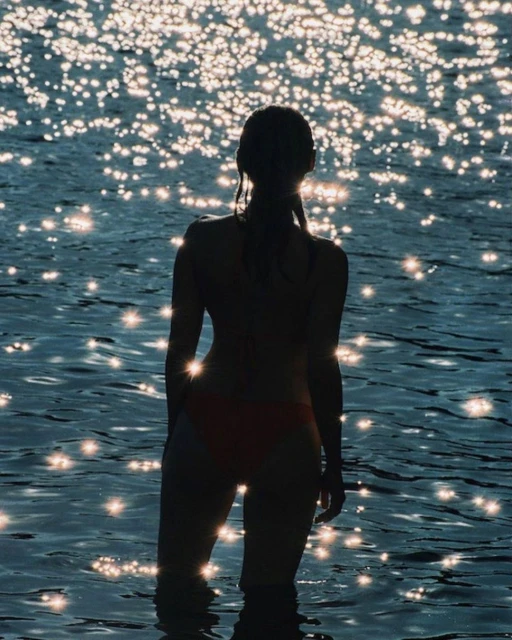 Silhouette of a young woman in reddish swimsuit standing in water at daytime, reflecting sunlight with sparkling effect in the water.