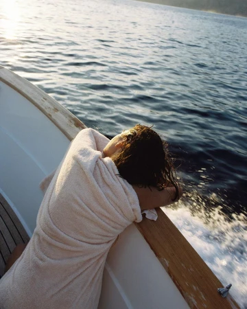Woman wrapped in a towel leaning on the wooden railing of a moving boat overlooking dark water