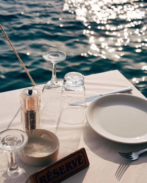 Table setting featuring wine glasses, plates, and cutlery on a white tablecloth, set against a sparkling ocean backdrop, creating an elegant ambiance.