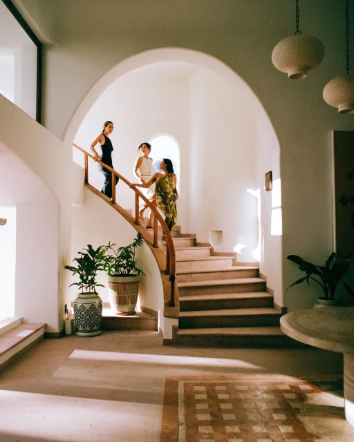 Three women are walking down a staircase indoors; two potted plants are visible on the left and hanging lights above.