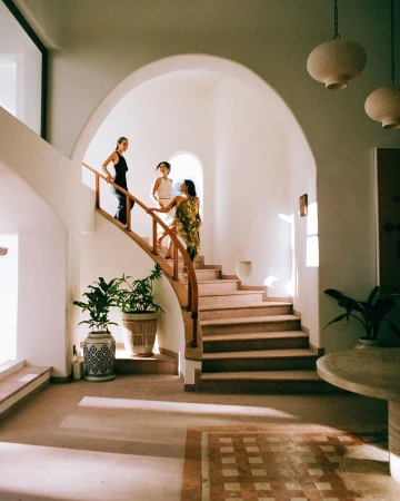 Three women are walking down a staircase indoors; two potted plants are visible on the left and hanging lights above.