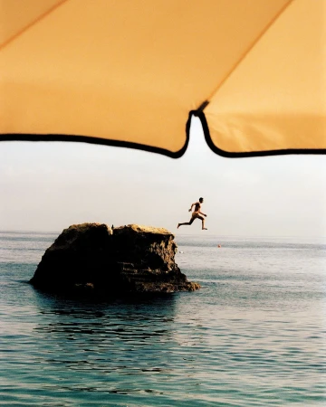 A man jumps from a rock into the ocean, seen from under a yellow beach umbrella, capturing a summer vacation moment.