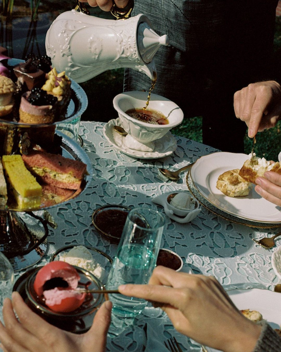 Afternoon tea scene: scones, cakes, tea being poured into a cup on a table with patterned tablecloth in an outdoor setting.
