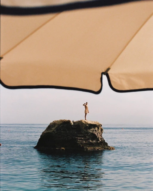 A female figure stands atop a rock formation in the ocean, under a beach umbrella, on a sunny day with clear skies and calm water.