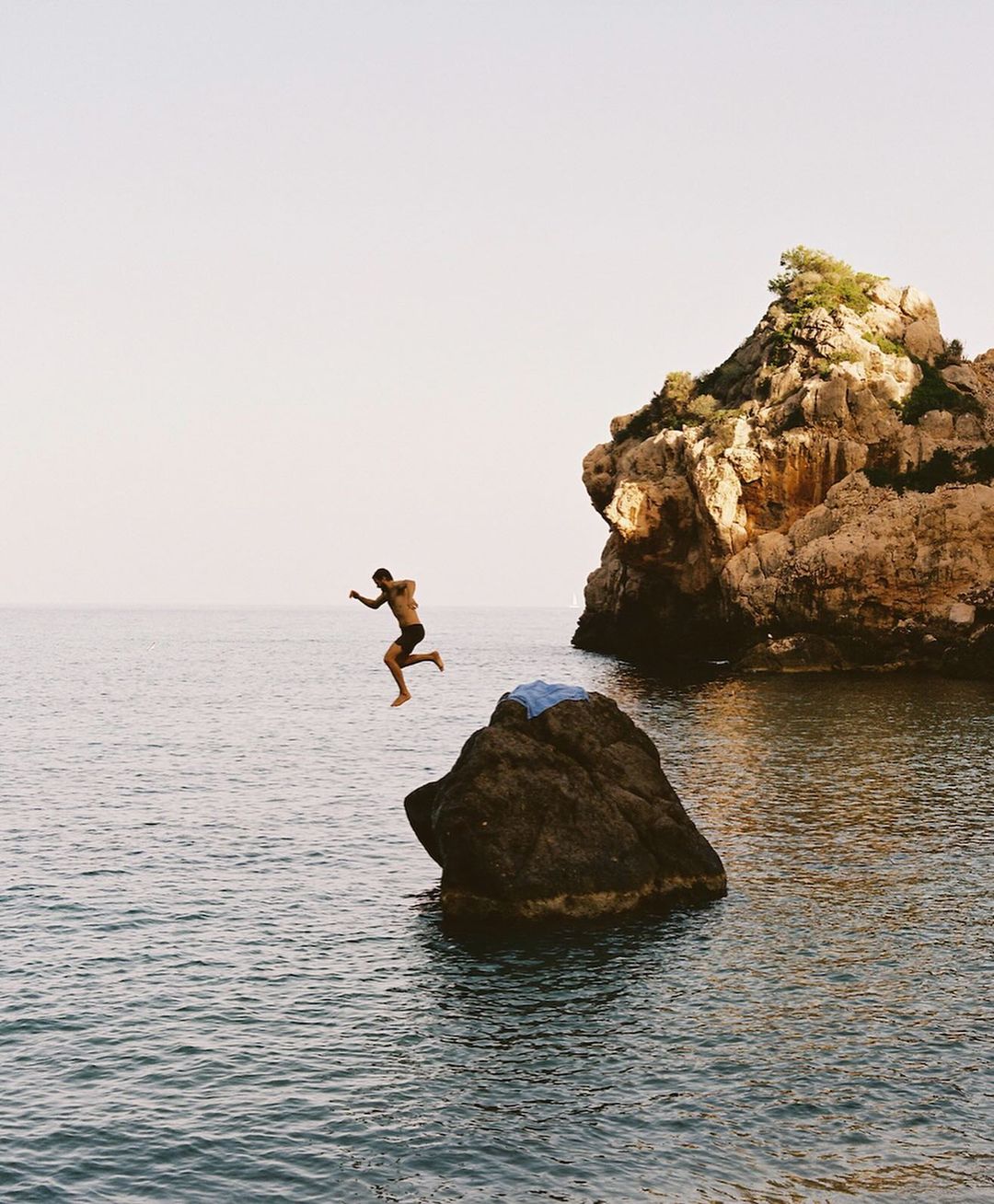 Man jumping off a rocky outcrop into the open sea under a clear sky.