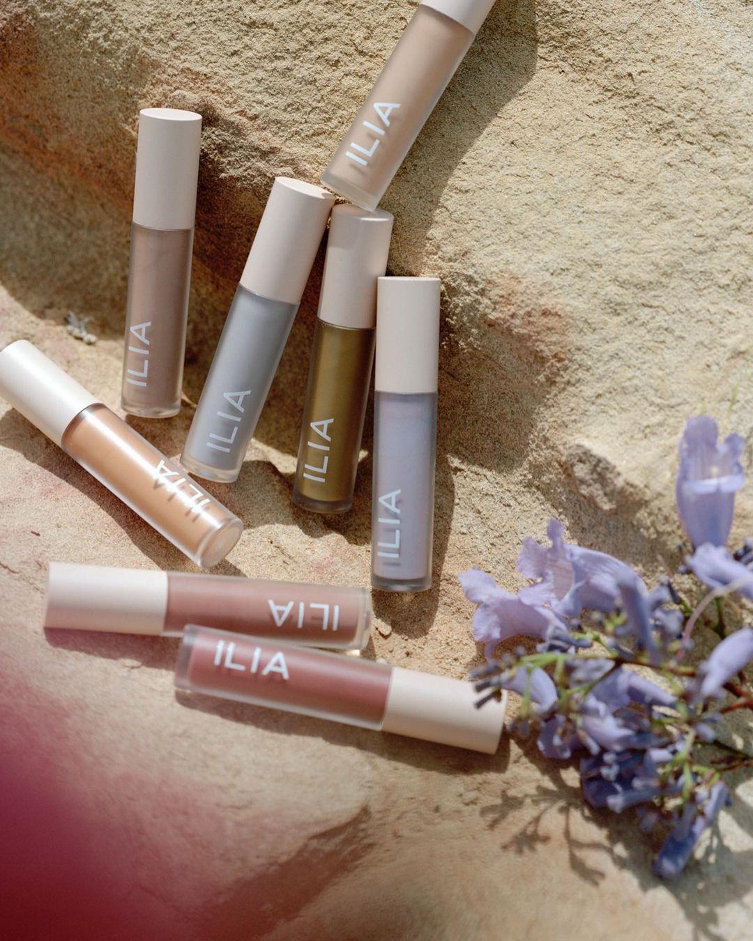 Several tubes of Ilia lipsticks and makeup displayed on a sandy surface with purple flowers, showcasing a beauty product arrangement.