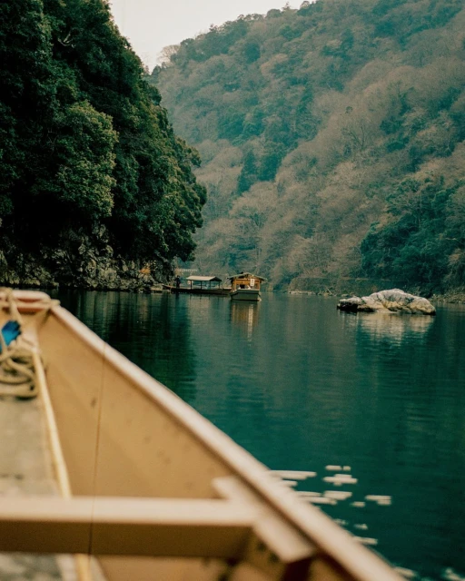 Calm lake scene with a wooden boat in the foreground and a river boat on the serene water, surrounded by lush, green mountains.