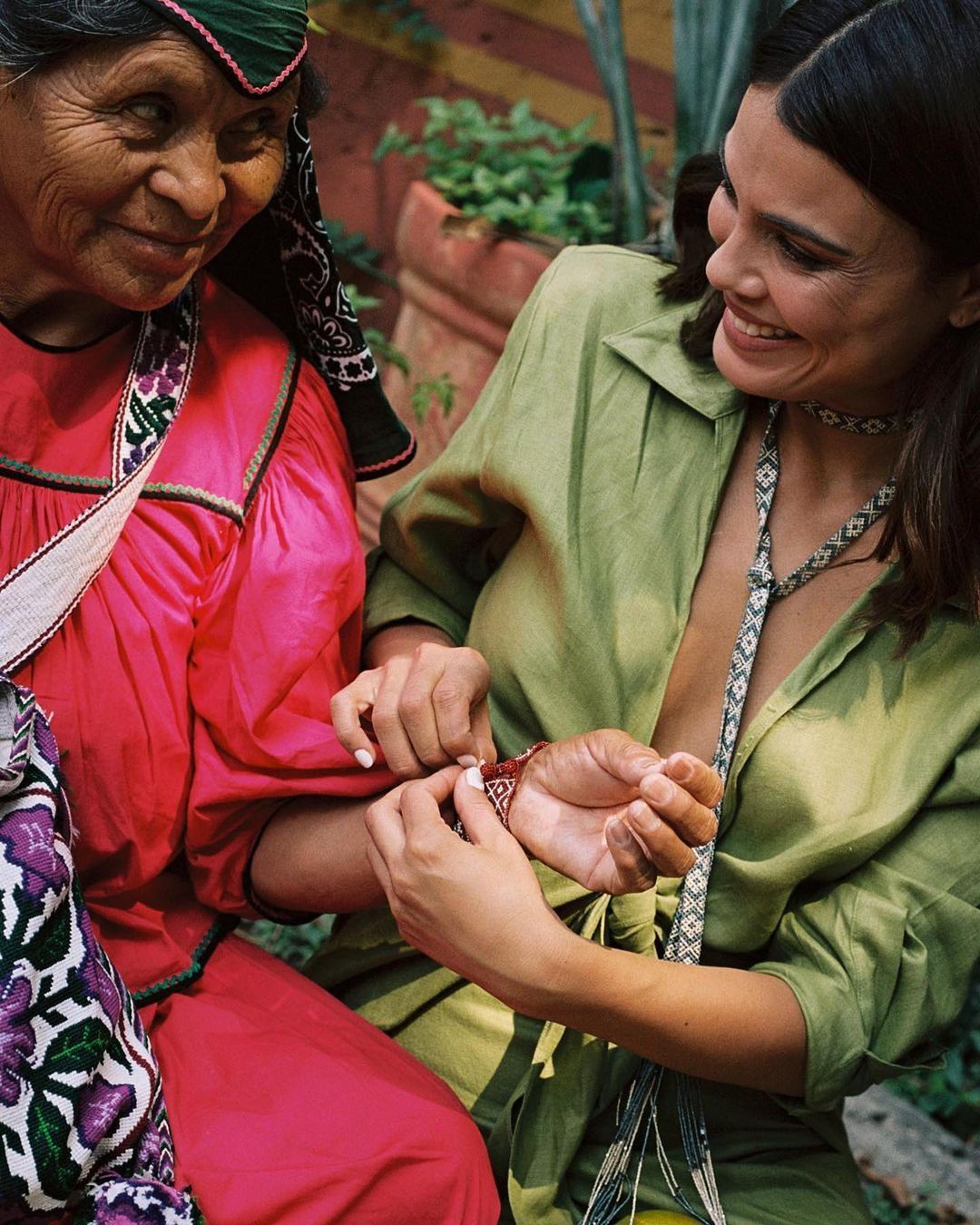 Two women smiling outdoors. One woman in red traditional dress with floral trim is being gifted a bracelet by a younger woman in green.