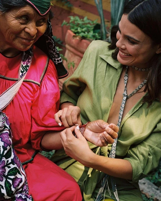 Two women smiling outdoors. One woman in red traditional dress with floral trim is being gifted a bracelet by a younger woman in green.