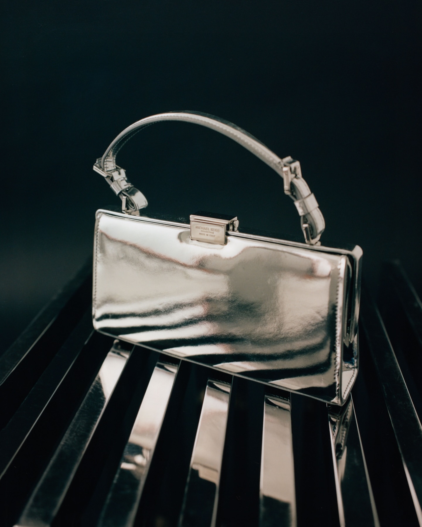 Silver rectangular clutch bag with a top handle displayed on a background of shiny silver bars, creating a striking, reflective texture.