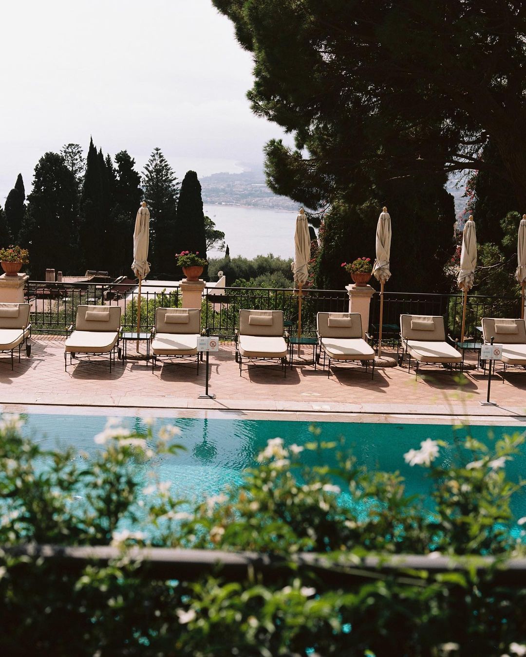Outdoor resort scene featuring a swimming pool lined with lounge chairs and umbrellas, overlooking a coastal landscape with greenery.