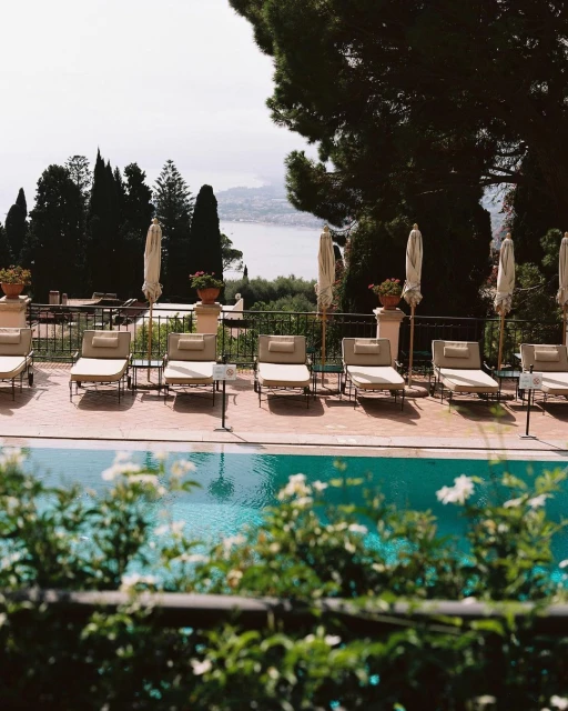 Outdoor resort scene featuring a swimming pool lined with lounge chairs and umbrellas, overlooking a coastal landscape with greenery.
