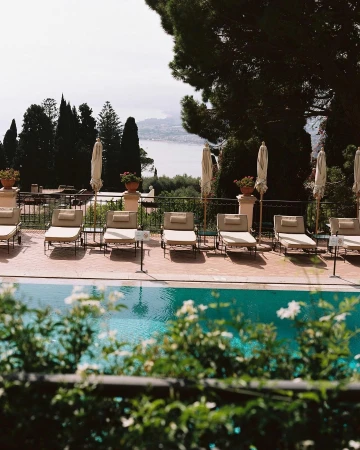 Outdoor resort scene featuring a swimming pool lined with lounge chairs and umbrellas, overlooking a coastal landscape with greenery.