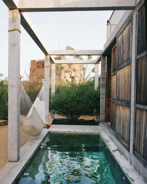 Outdoor resort scene showcases a white hammock strung between concrete pillars, reflecting in a tranquil pool, with rustic architecture.